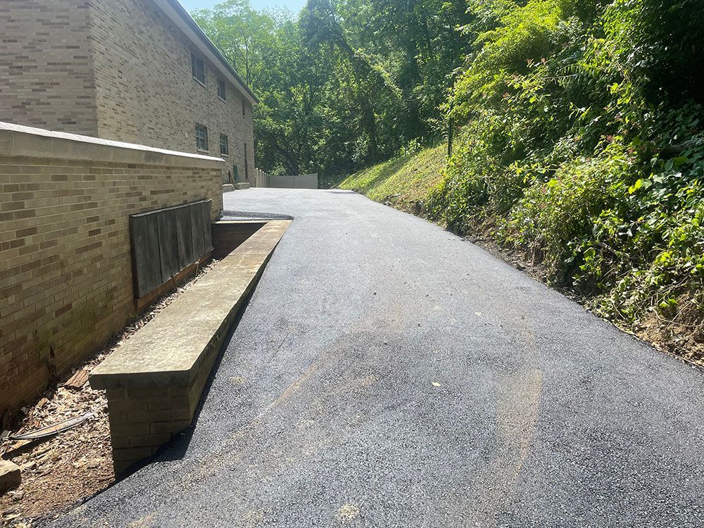 Asphalt driveway next to a brick building and a wooded hillside.