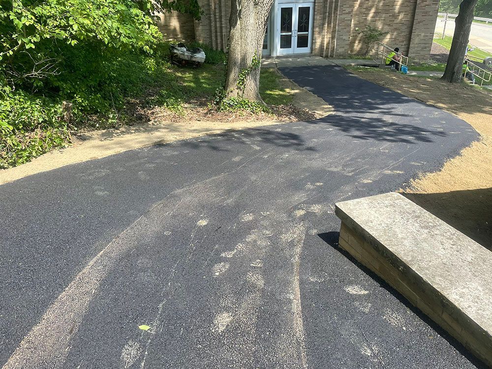 Newly paved asphalt driveway leading to a building's entrance. Trees and a concrete ledge are visible.