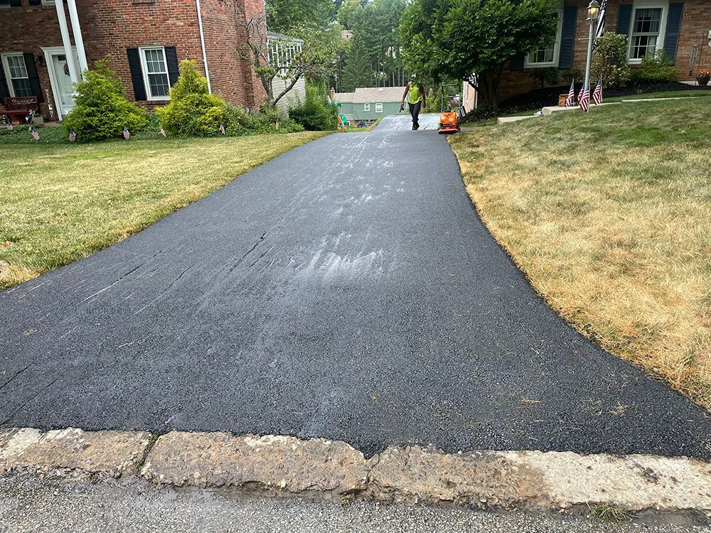 Newly paved asphalt driveway with person operating a machine; flanked by lawns, brick buildings.