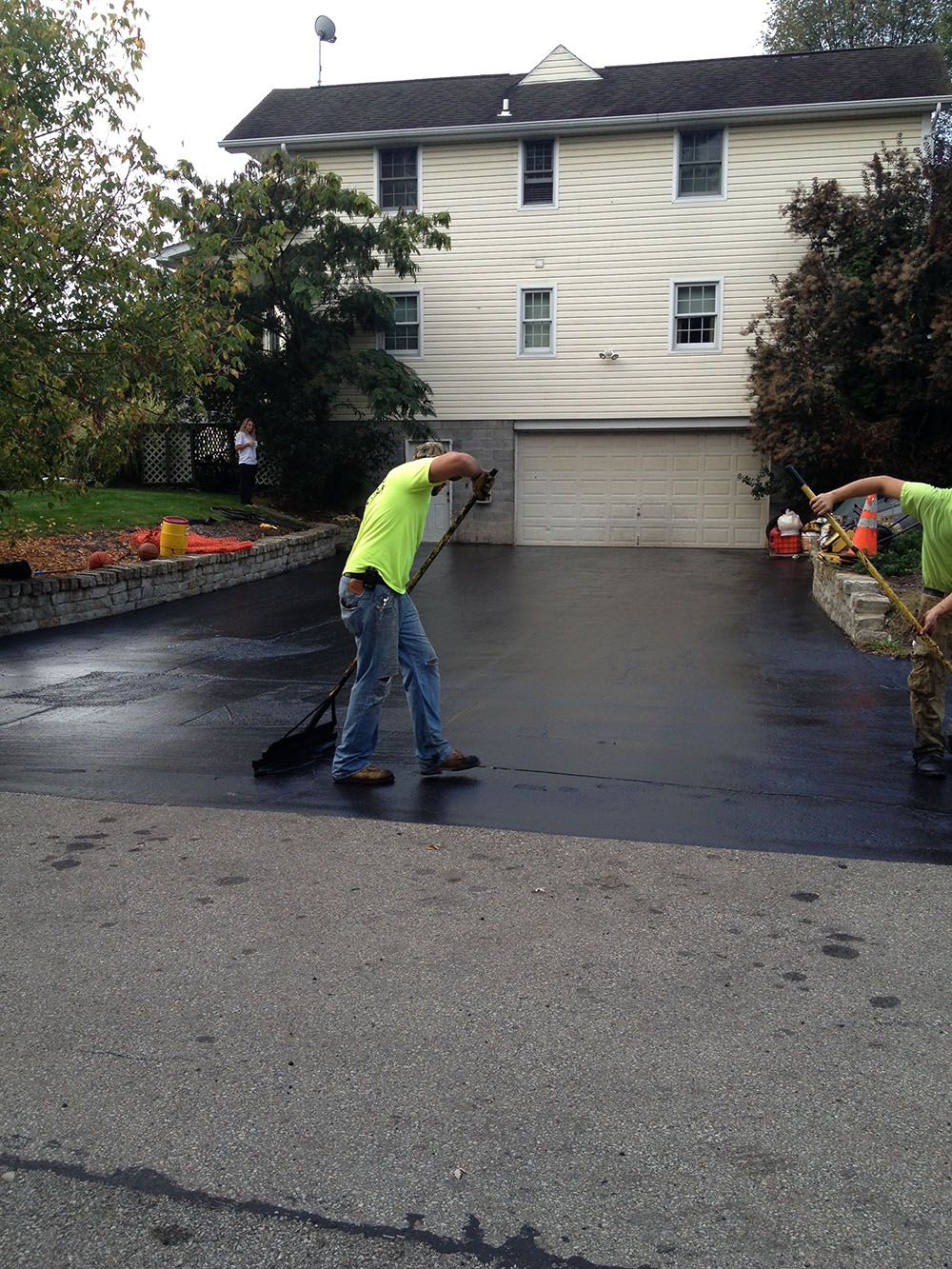 Two workers in green shirts paving a driveway. One sweeps, the other gestures. House in background.