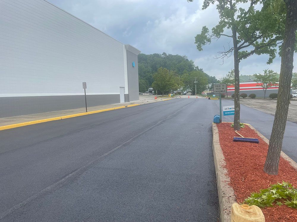 Black asphalt road next to a white building and a landscaped area with mulch and trees.