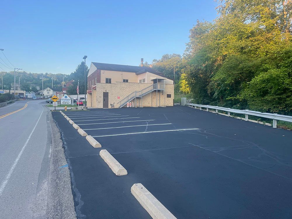 Paved parking lot in front of a tan two-story building; road on the left, trees on the right, under a blue sky.