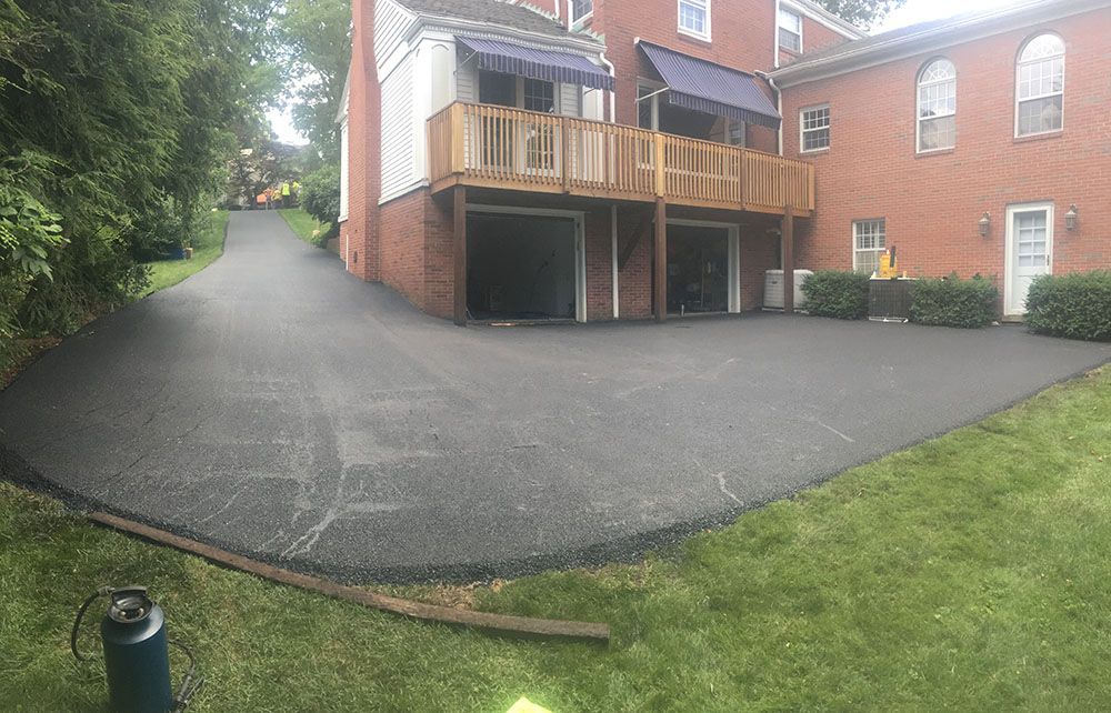 Asphalt driveway leading to a brick house with a wooden deck and two garage doors; lush green grass surrounds.