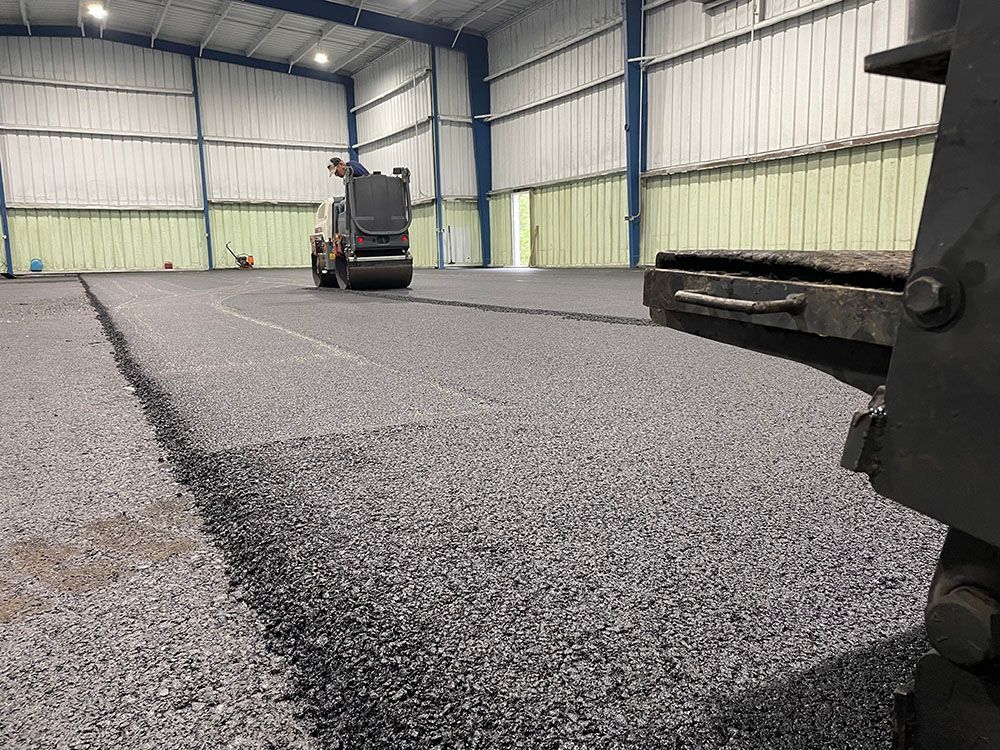 Asphalt being laid inside a large metal building. A worker operates a roller.