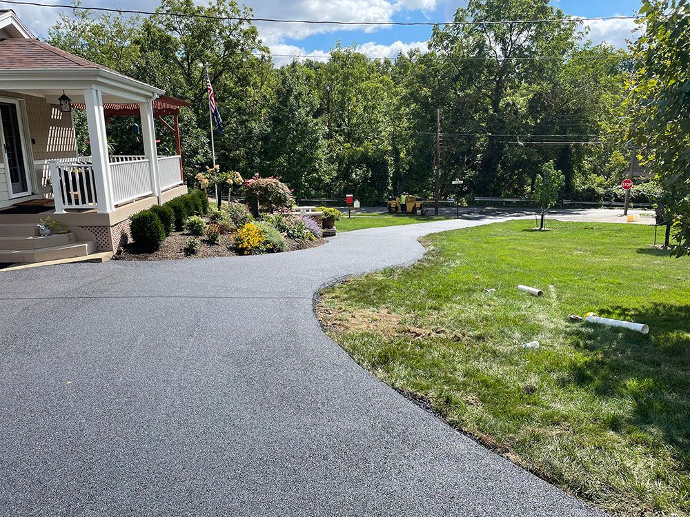 A paved, dark driveway curves towards a house with a porch. Landscaping and grass border the driveway.