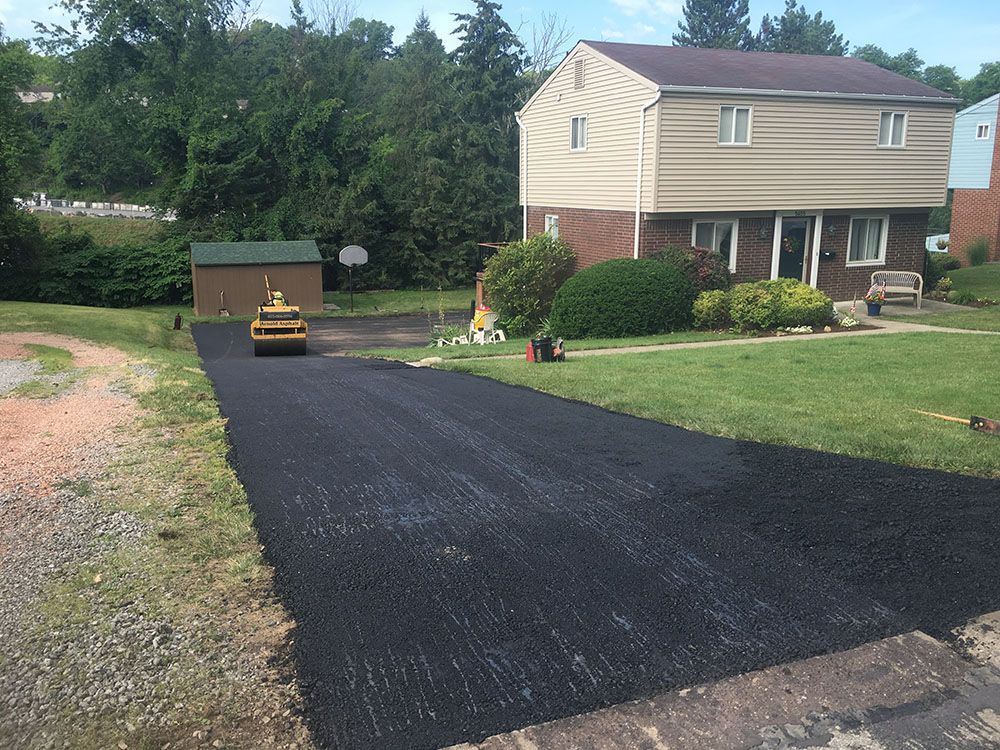 Asphalt driveway being rolled by a compactor in front of a two-story beige house and green lawn.