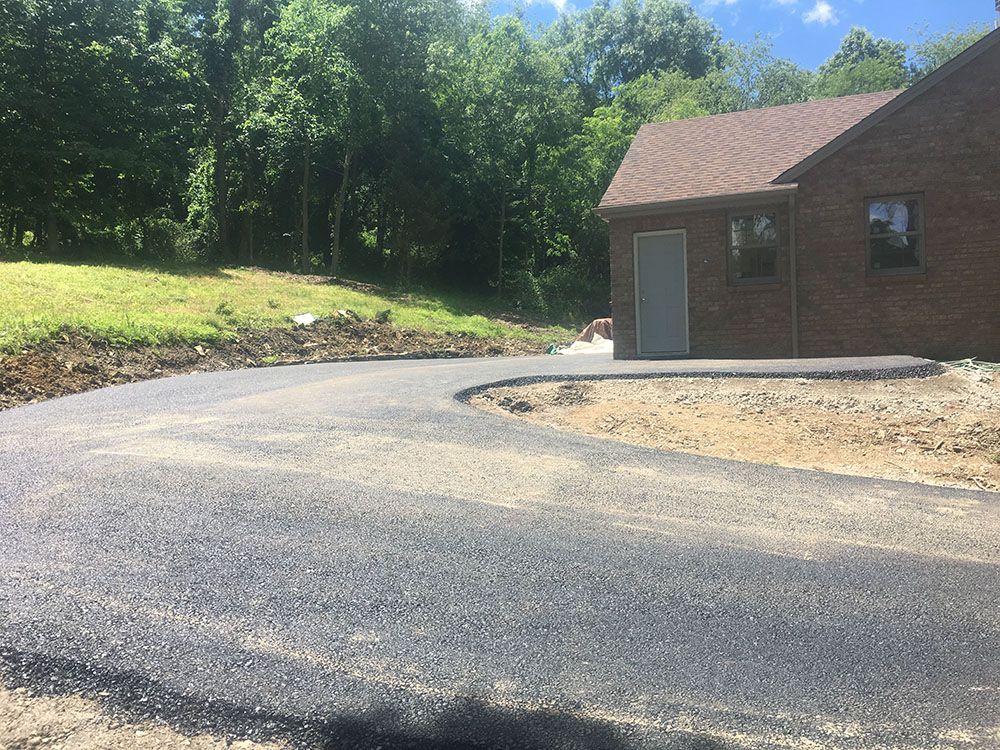 Asphalt driveway curving towards a brick building with a door and brown roof, set in a grassy area with trees.