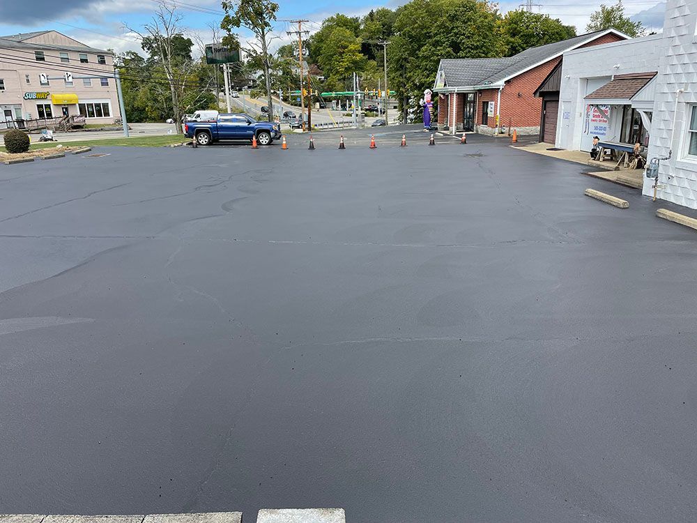 Newly paved asphalt parking lot with buildings in the background. A blue pickup truck and traffic cones are present.