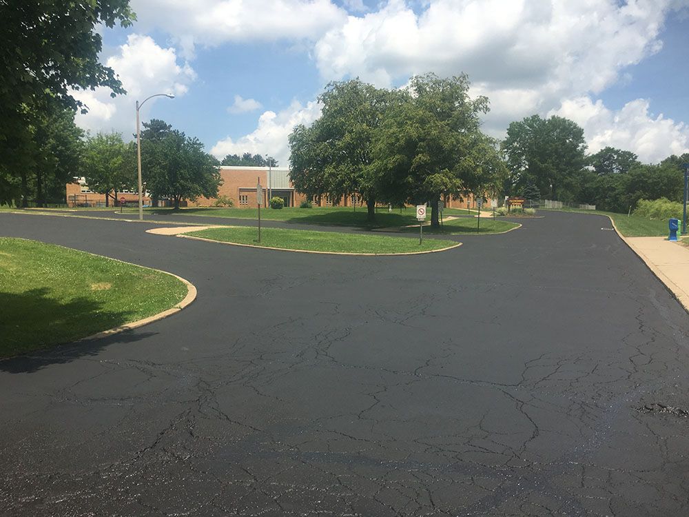 Newly paved asphalt road curves in front of a low brick building under a sunny sky.