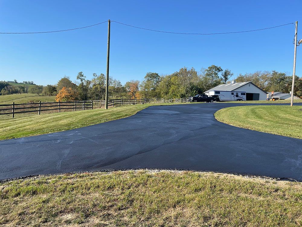 Paved driveway curves toward a white building, under a blue sky, with surrounding green grass and a wooden fence.