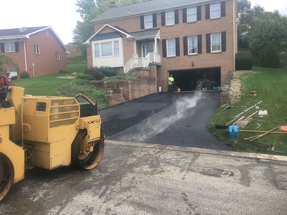 Yellow asphalt roller compacting freshly paved driveway in front of a brick house.