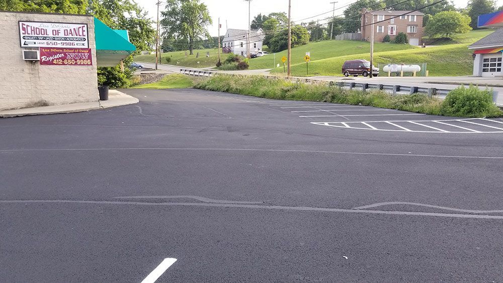 Paved parking lot with a building on the left, grassy area, and a road. Car in the distance.