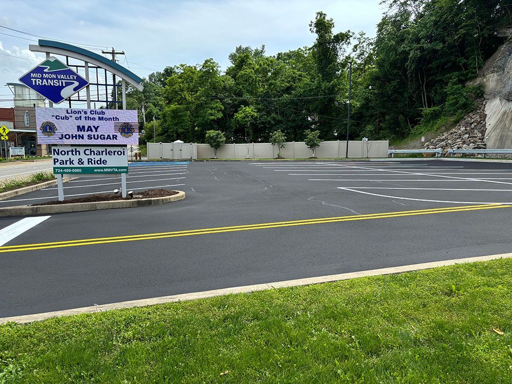 New asphalt parking area with sign for North Charleroi Park & Ride, next to a hill and trees.