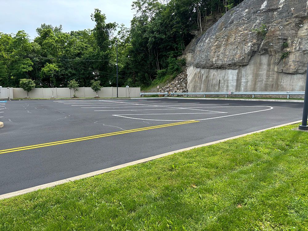 Asphalt parking lot next to a grassy area and rocky hillside with trees.