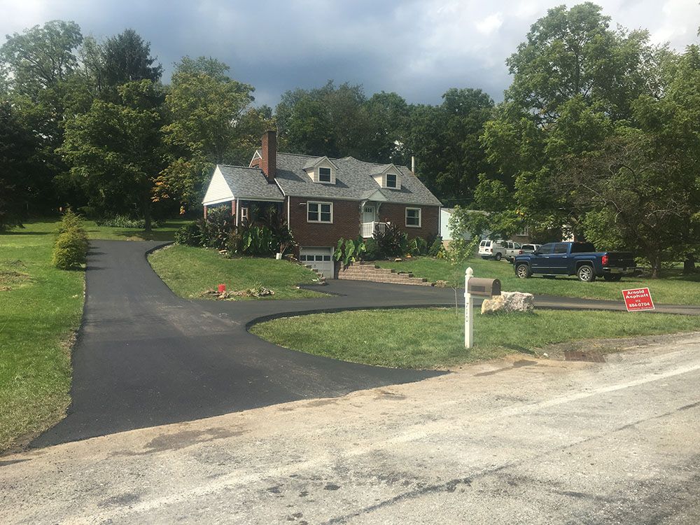Paved driveway leads to a brick house with a green lawn. Trees and a cloudy sky are in the background.