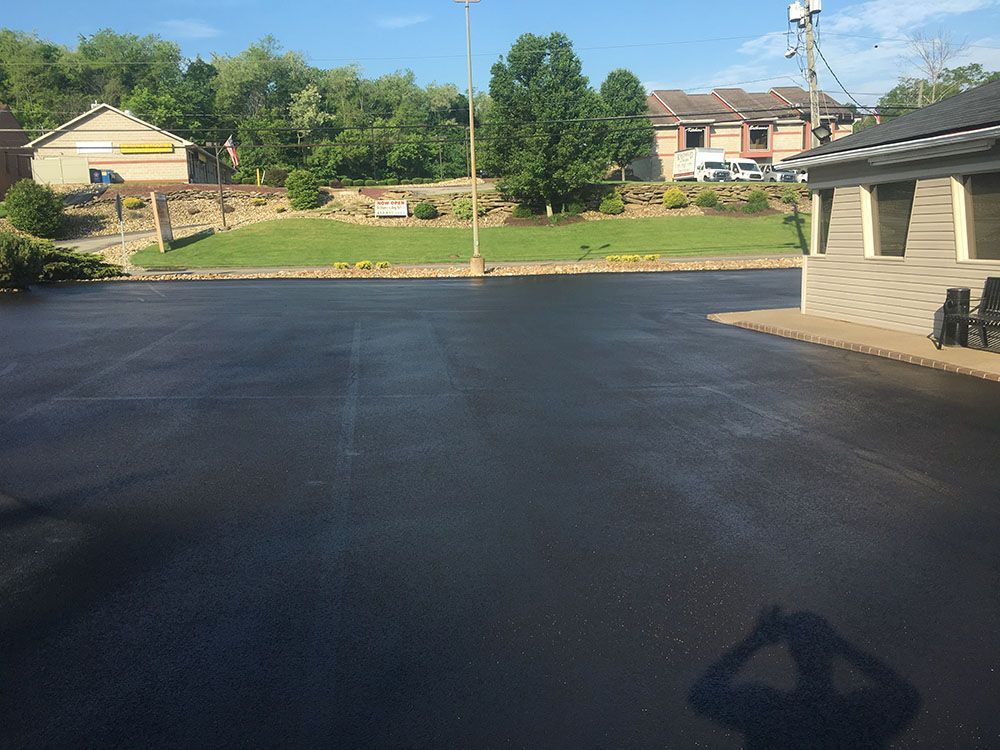 Freshly sealed black asphalt parking lot in front of buildings with green grass and trees.