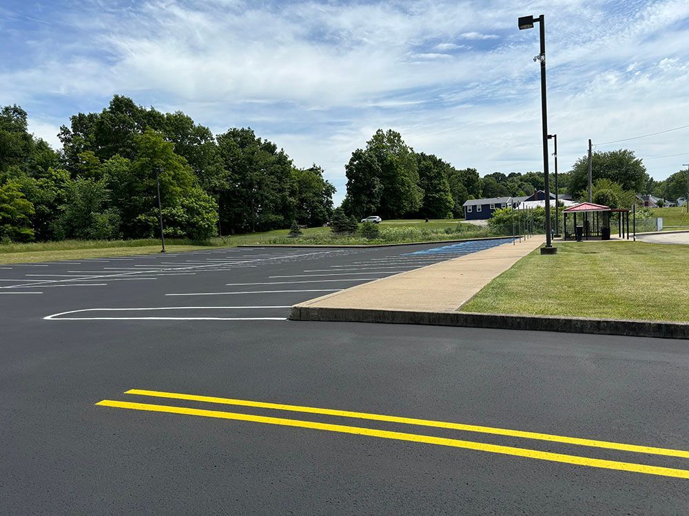 Asphalt parking lot with sidewalk, trees, and street lights under a partly cloudy sky.