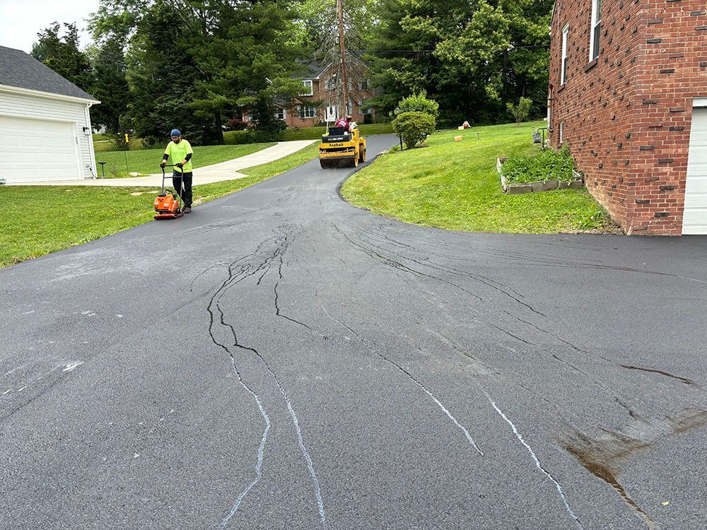 Driveway with cracks being repaired by a worker using an asphalt saw; a skid steer is also present.
