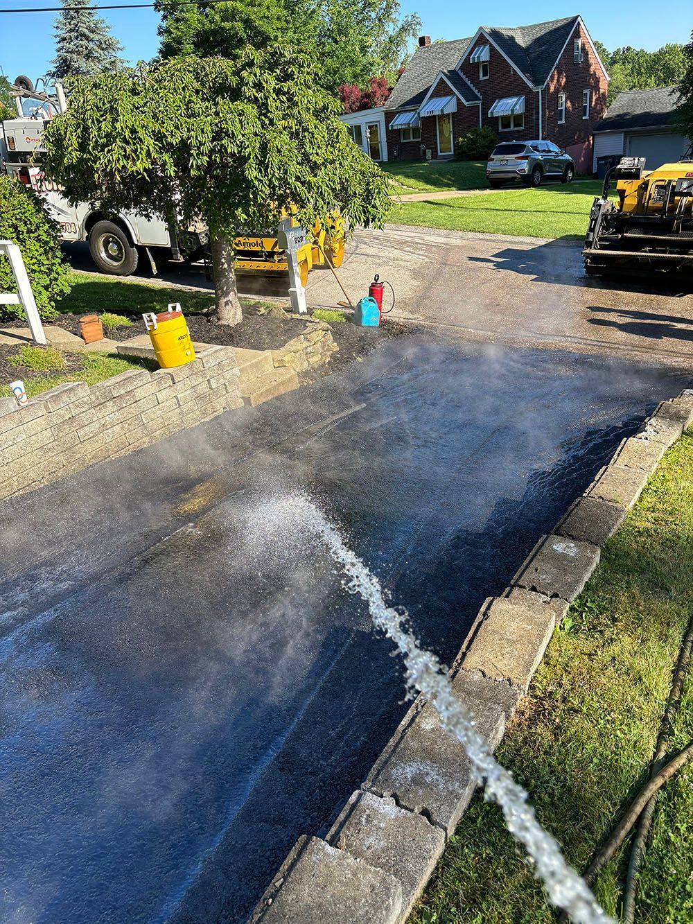 A black asphalt driveway being sprayed with water; a maintenance vehicle and houses in the background.