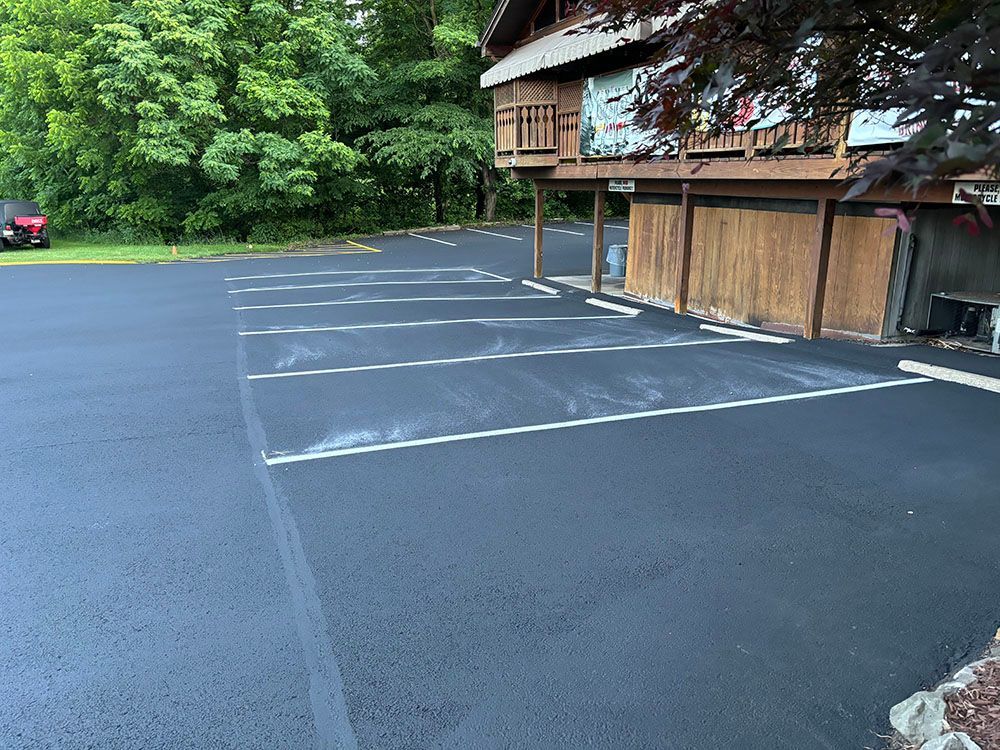 Black asphalt parking lot with white-lined spaces, next to a wood-paneled building with a porch, trees in the background.