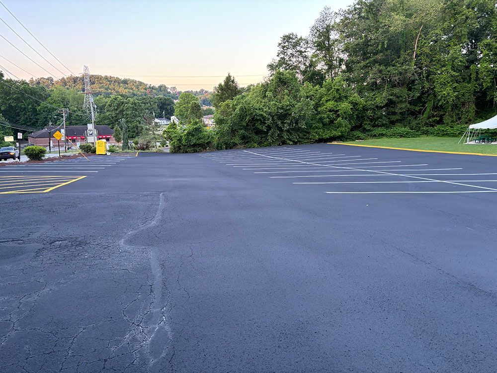 Empty, freshly paved parking lot with painted lines, backed by trees and a hillside. Buildings visible at left.
