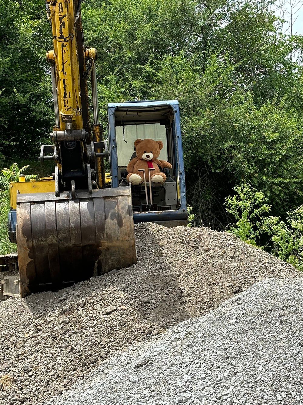 Teddy bear sitting in the cab of a yellow and blue excavator, parked on a pile of gravel.