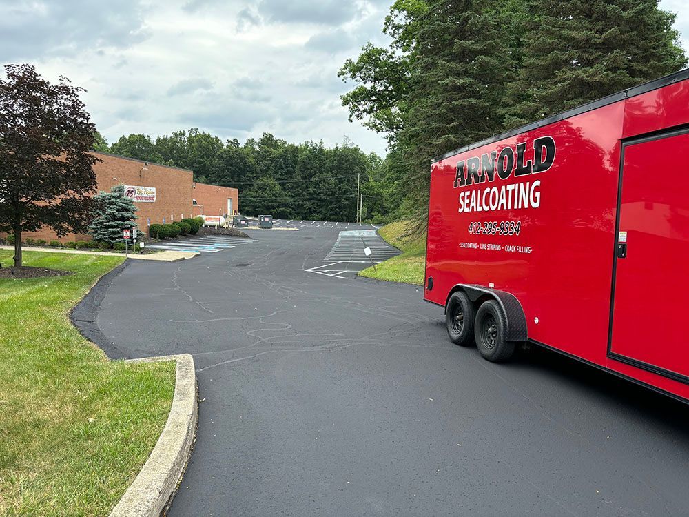 Red trailer with Arnold Sealcoating parked on a newly sealed asphalt driveway, next to a building.
