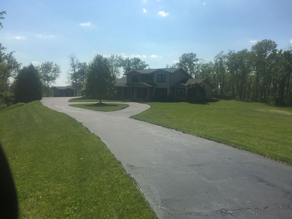 A long, winding asphalt driveway leads to a two-story house surrounded by green grass and trees under a blue sky.