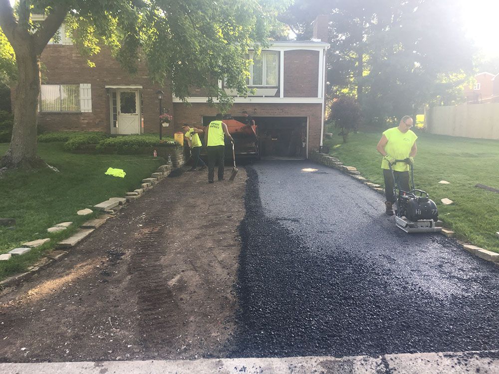Asphalt driveway being paved; workers in safety vests; one using a compactor.