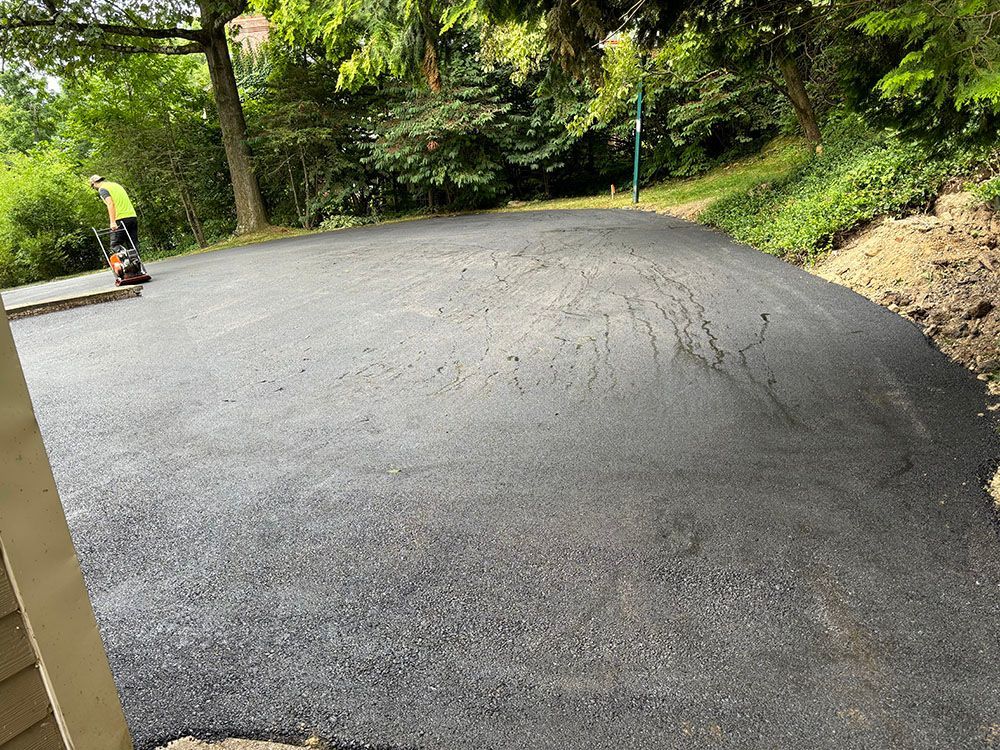Person operating a machine on a newly paved asphalt surface, likely a driveway. Green trees surround.
