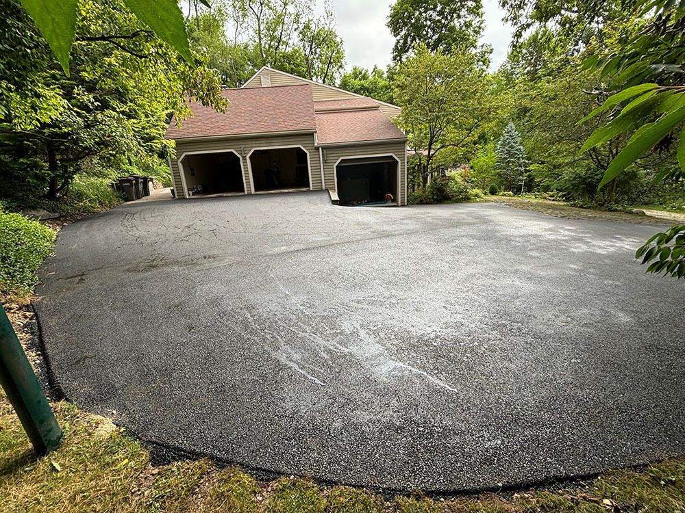 Asphalt driveway leading to a three-car garage in a wooded setting. The garage has a brown roof.