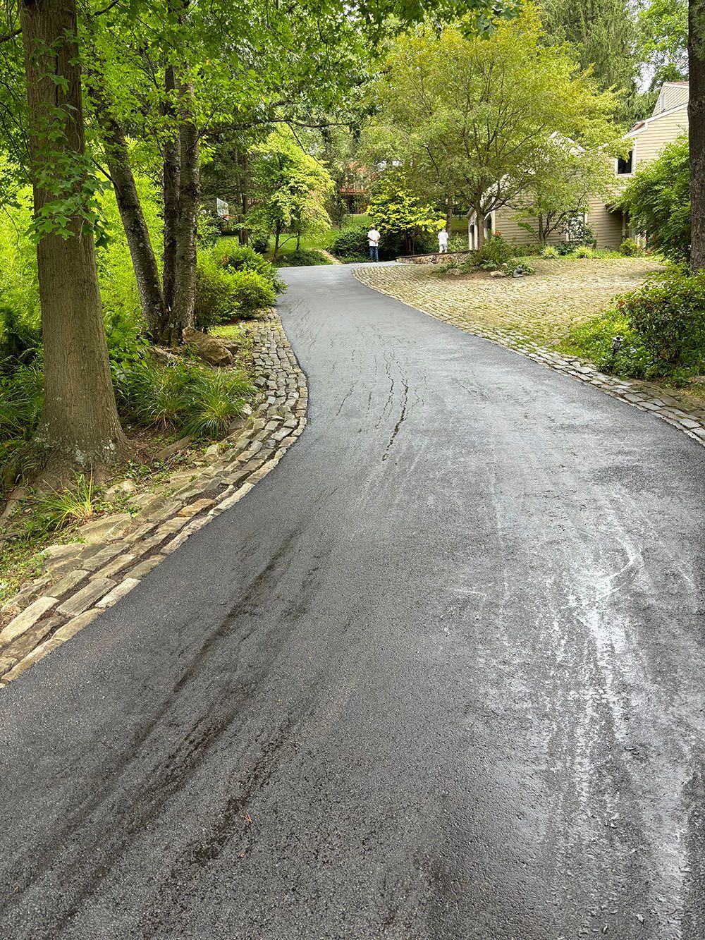Asphalt driveway curves toward a house, bordered by trees and stone retaining wall.