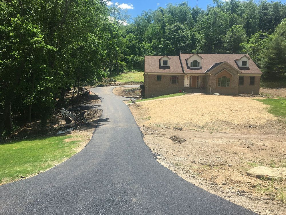 Asphalt driveway leading to a brick house with tan landscaping. Trees surround the property.