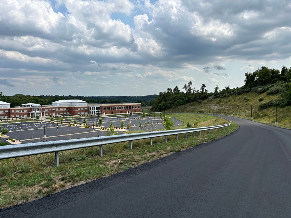 Paved road curves towards a large school building and expansive parking lot under a cloudy sky.