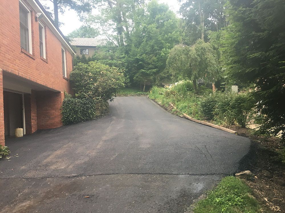 Asphalt driveway leading uphill, beside a brick house and greenery.