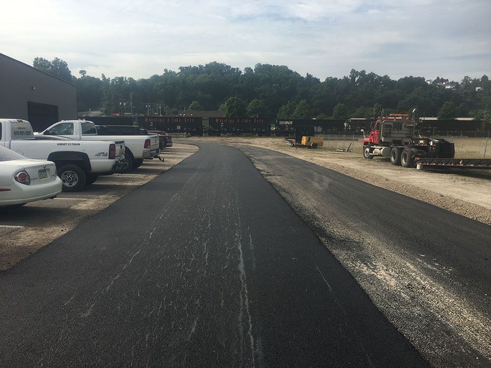 Newly paved asphalt road; parked trucks and a building on the left, trees in the background.