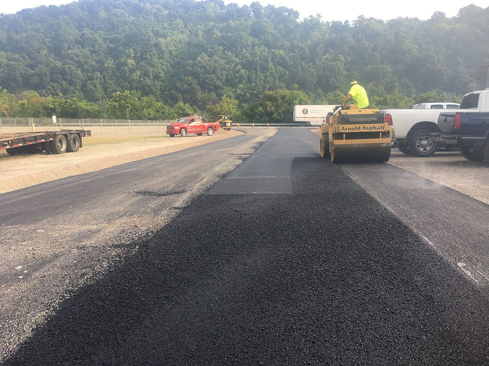 Asphalt paving on road, construction worker operates roller, vehicles parked nearby, mountains in the background.