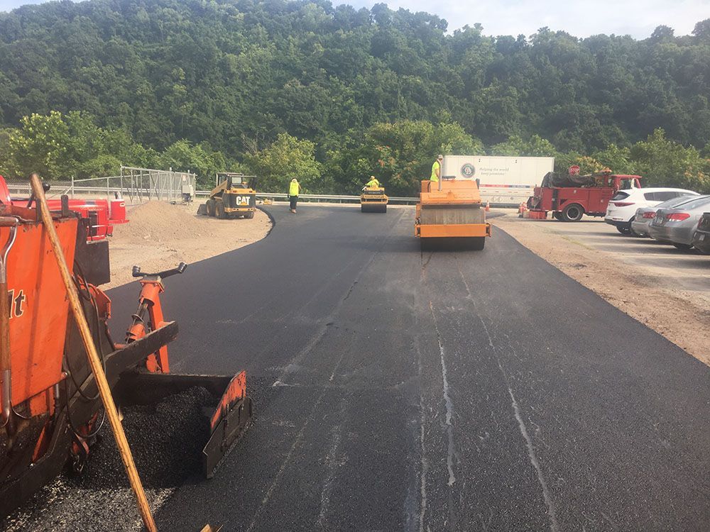 Asphalt paving in progress on a road, with machinery, workers, and a forested backdrop.