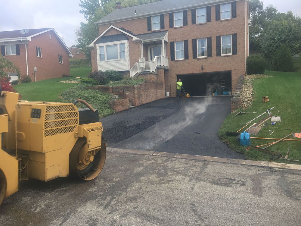 Yellow asphalt roller compacting freshly paved driveway in front of a two-story brick house.