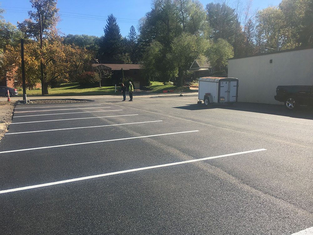 Newly paved parking lot with white lines. Two people stand near a trailer. Building in the background.