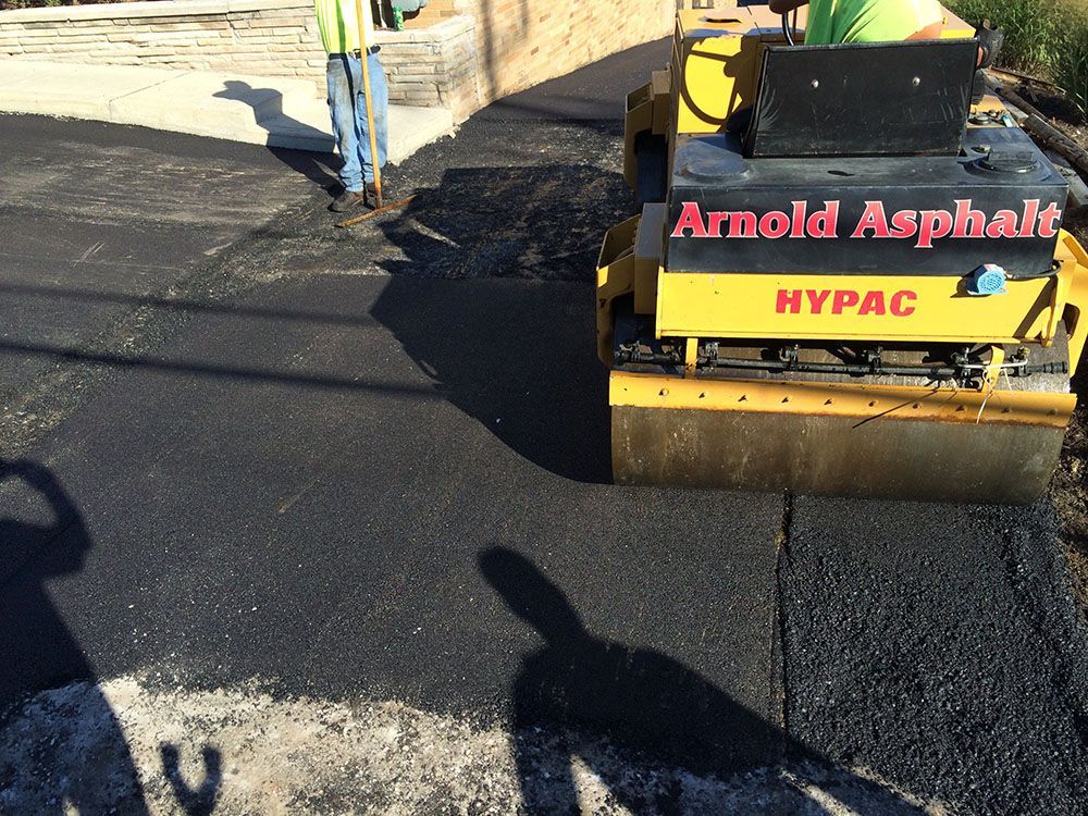 Asphalt roller compacting fresh pavement. A worker stands nearby.