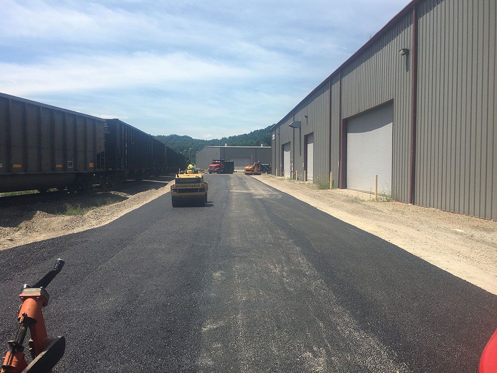 Asphalt road construction with a roller, train cars, and industrial building.