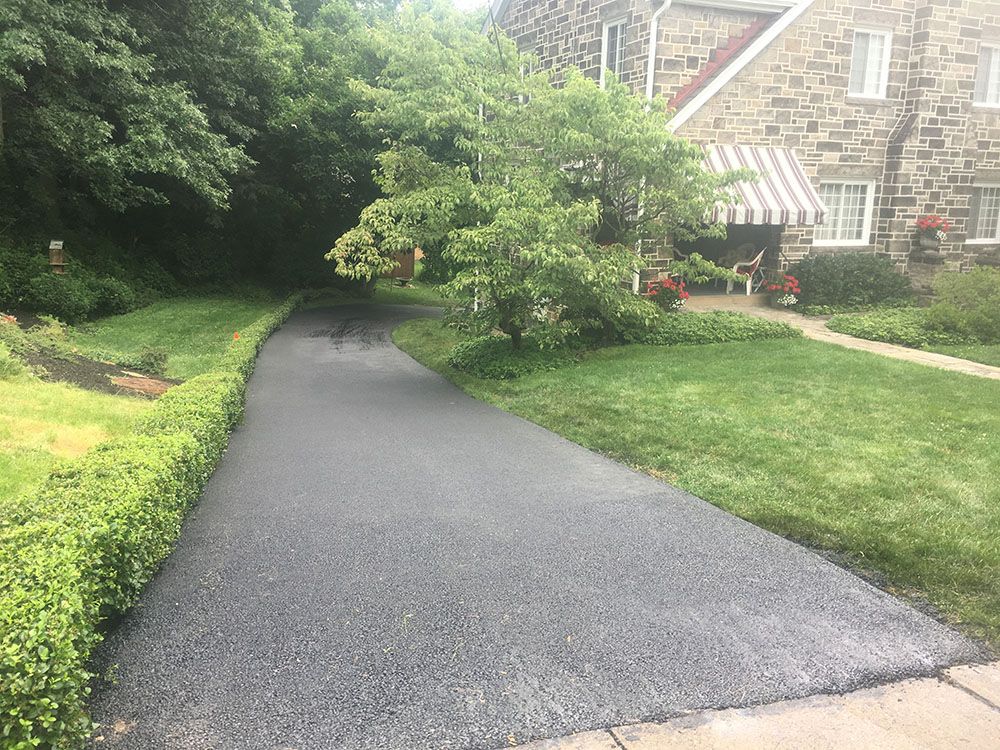 Paved driveway leading to a stone house with a green lawn. Trees line the left side.