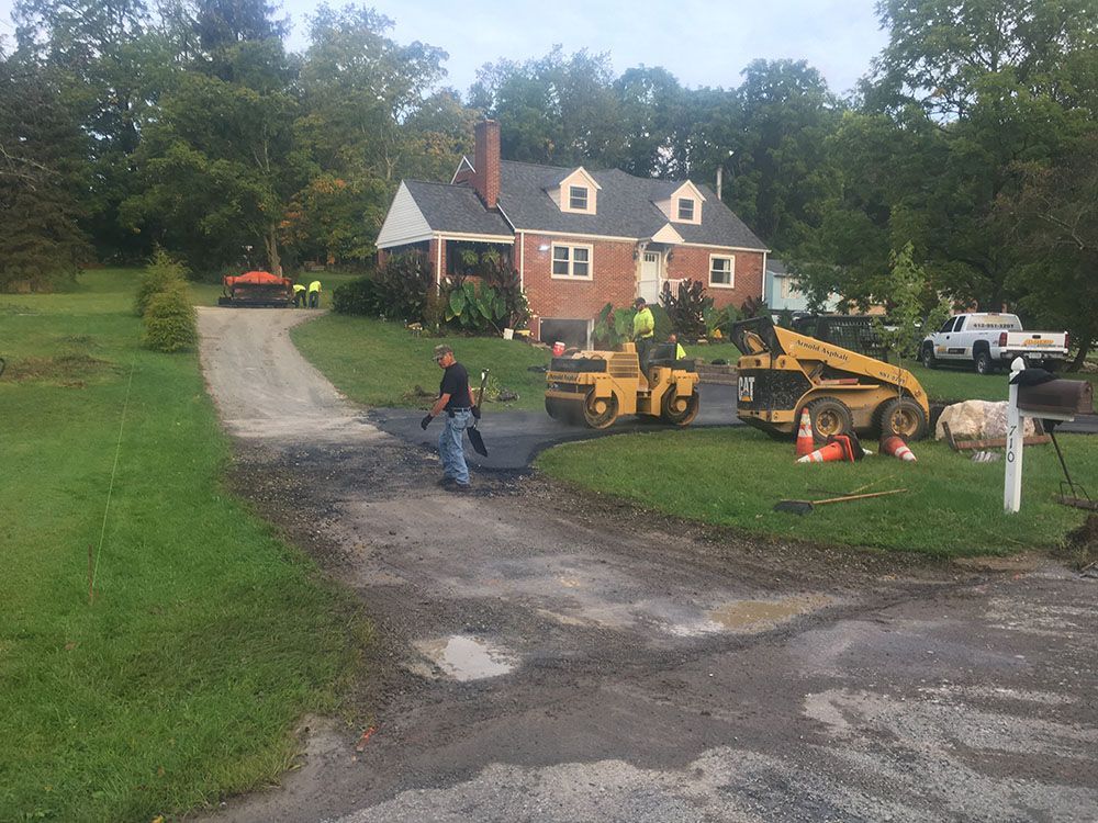 Driveway paving in progress at a brick house. Workers and machinery on site. Asphalt, grass, and trees.