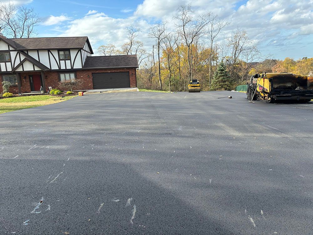 Newly paved asphalt driveway in front of a house with a Tudor-style facade; construction equipment present.