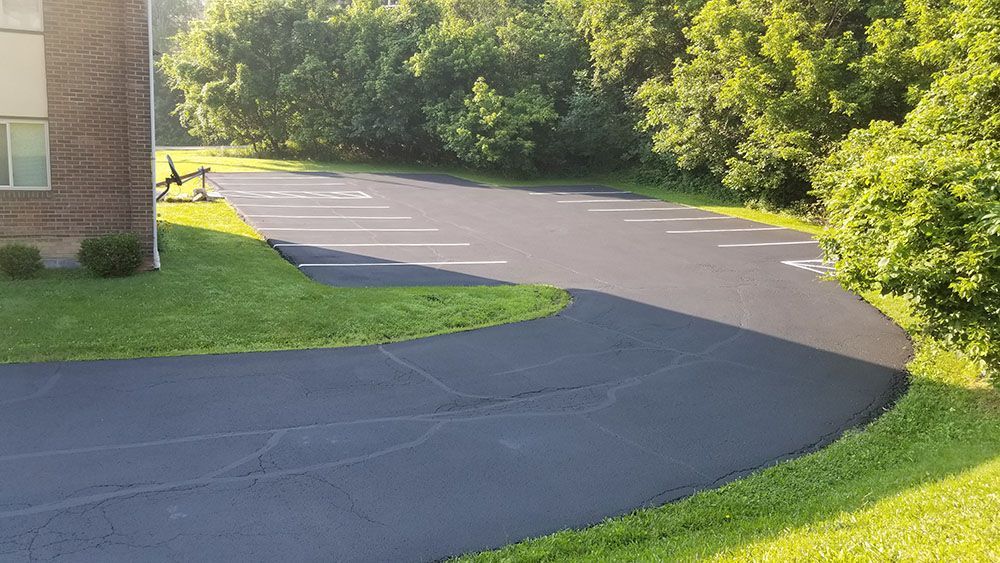 Asphalt parking lot with painted spaces next to a brick building and green grass, with trees in the background.