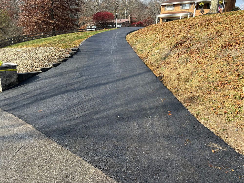 A newly paved asphalt driveway curves uphill towards a house on a sunny day.
