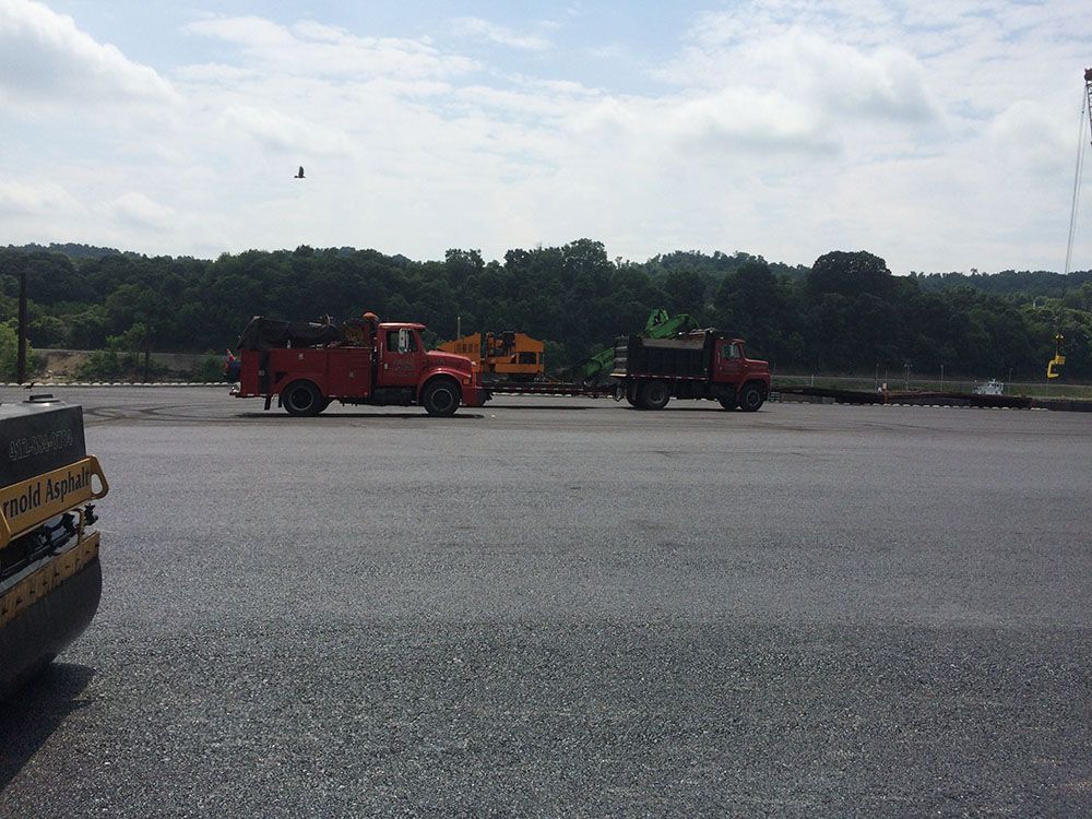 Construction site with two red trucks, a dark truck, and a roller on asphalt. Trees and sky in the background.