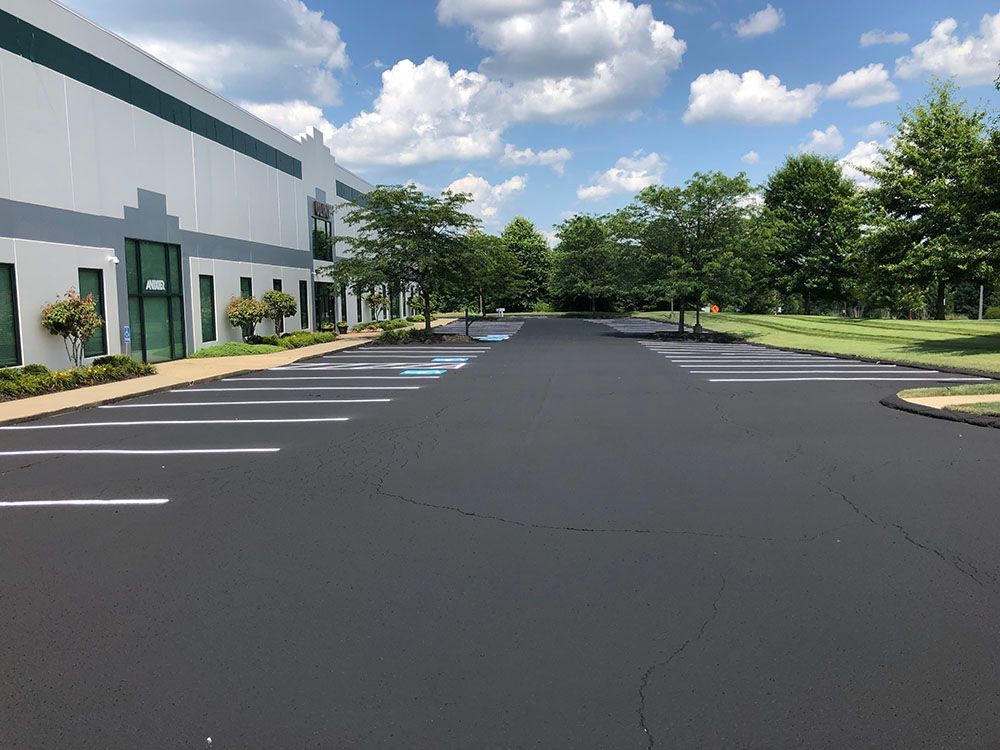 Black asphalt parking lot with white parking space lines next to a light-colored industrial building on a sunny day.