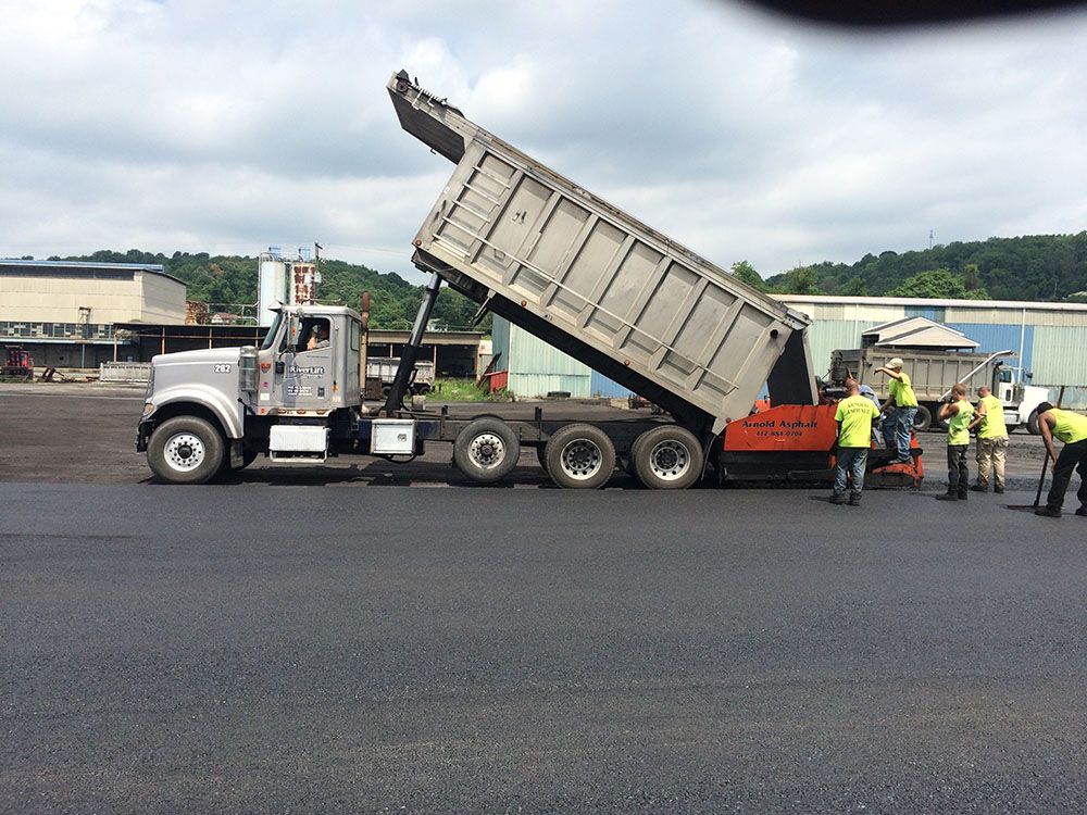 Dump truck unloading asphalt to feed a paving machine on a road. Workers in vests assist.